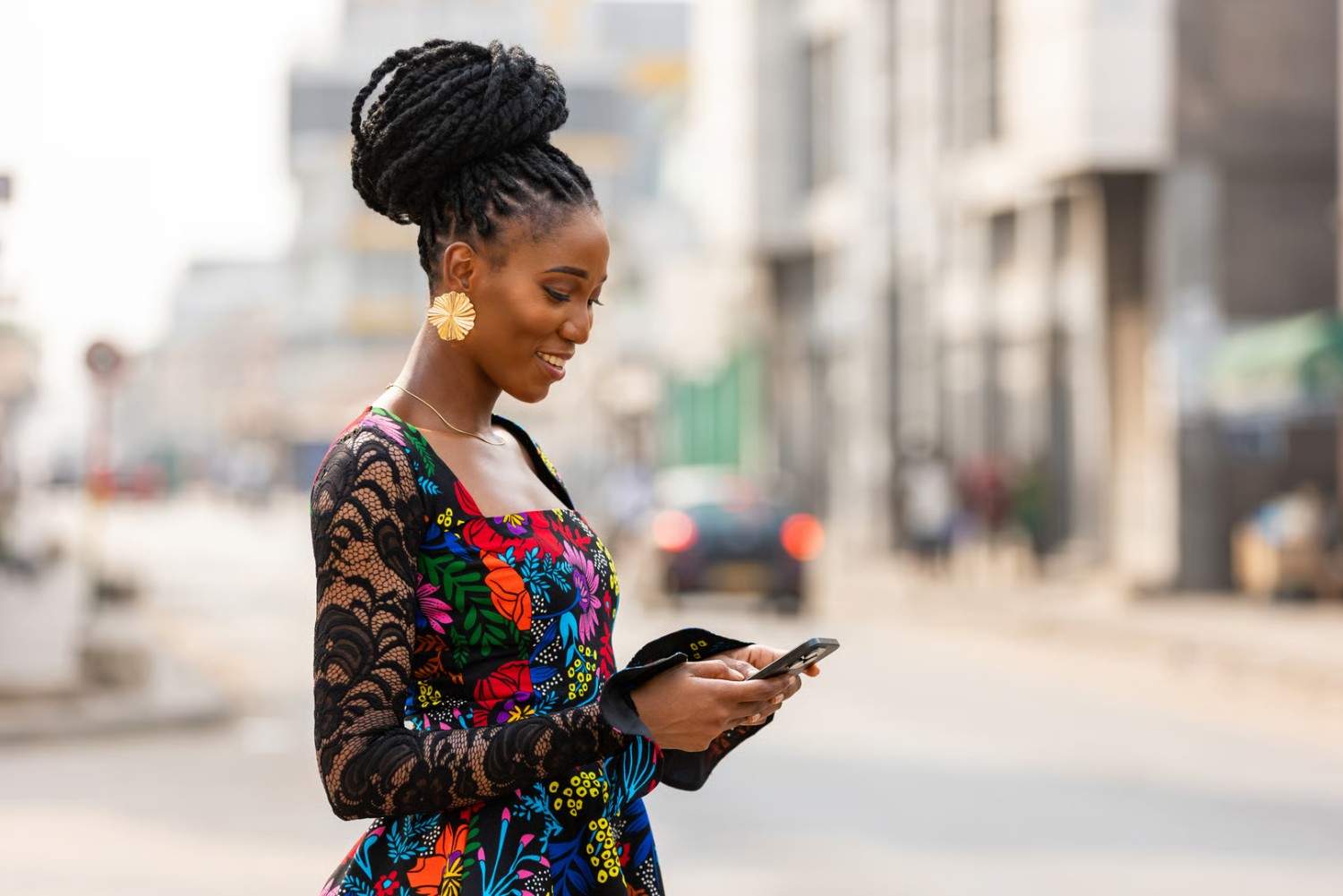 A fashionable African woman in a vibrant dress checks her phone outdoors. The urban setting is blurred, focusing attention on her.